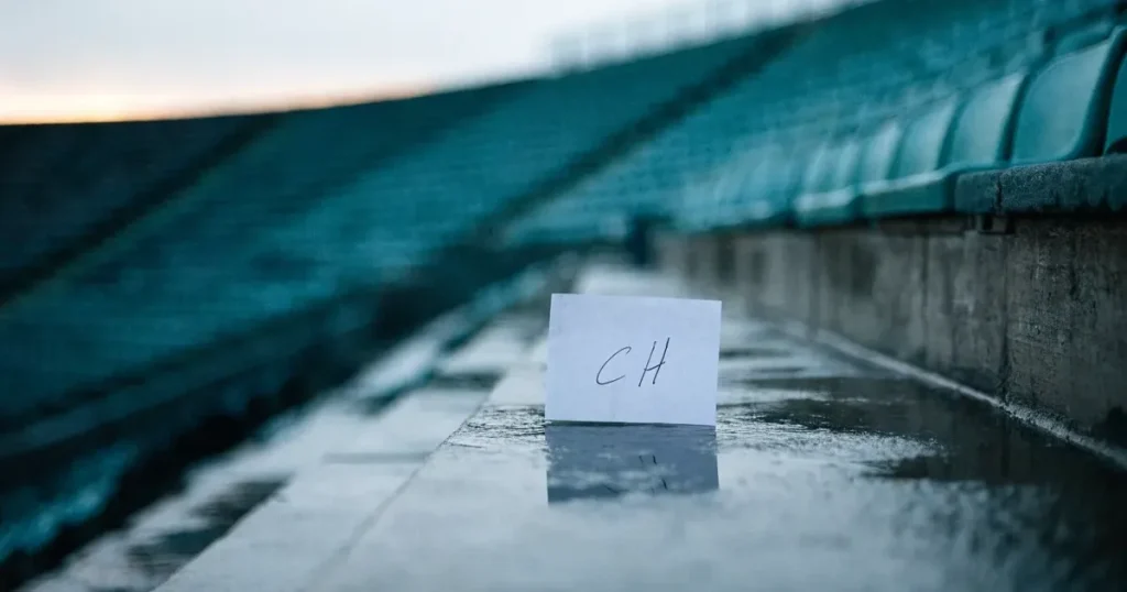 Eine Reihe leerer Sitzschalen auf der Tribüne eines kleinen Stadions, auf den Stufen glänzt Regenwasser.
