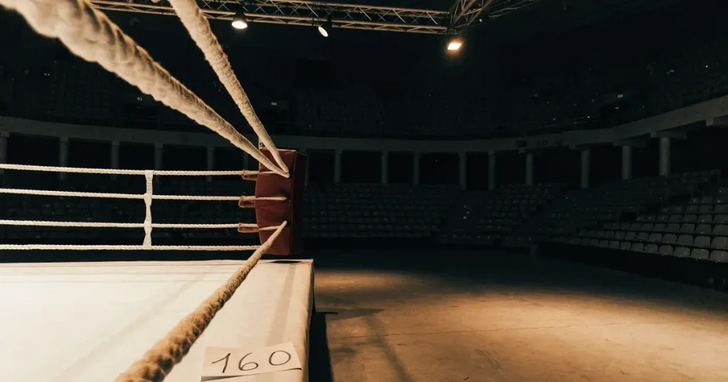Ein leerer Boxring mit gespannten Seilen unter hellem Hallenlicht, dahinter ansteigende Zuschauerränge in einer grossen dunklen Halle
