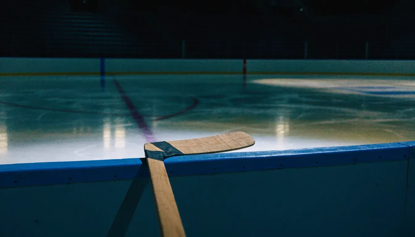 Ein Eishockeyschläger lehnt an der Bande, in der Ferne liegt die hell beleuchtete Eisfläche einer Halle unter Flutlicht.
