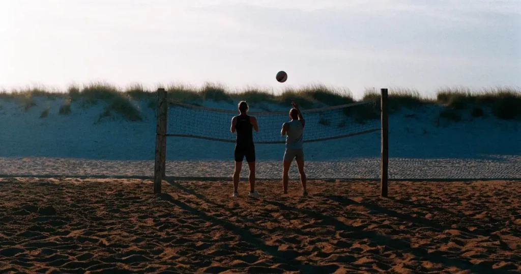 Ein Volleyball liegt im Sand eines Beachplatzes, zwischen zwei Pfosten ist ein Netz gespannt, tiefe Abendsonne wirft lange Schatten.