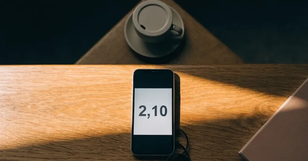 Ein Smartphone mit dunklem Display liegt auf einem Holztisch neben einem Ladekabel und einer Kaffeetasse.